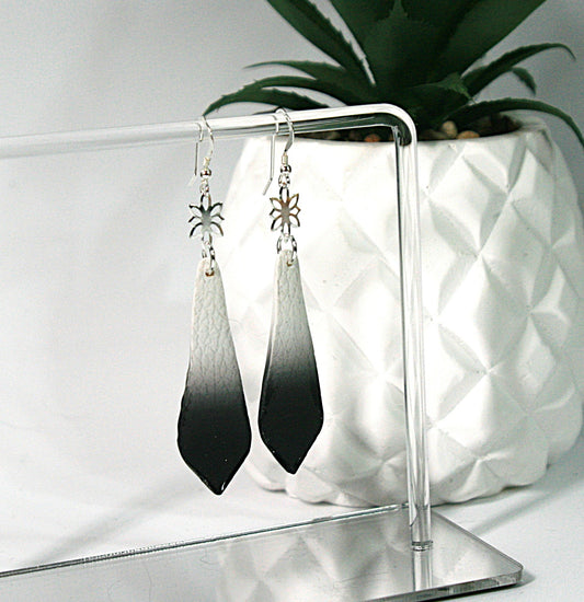 Black and white gradient earrings on a clear stand with a white textured surface and plant in the background.